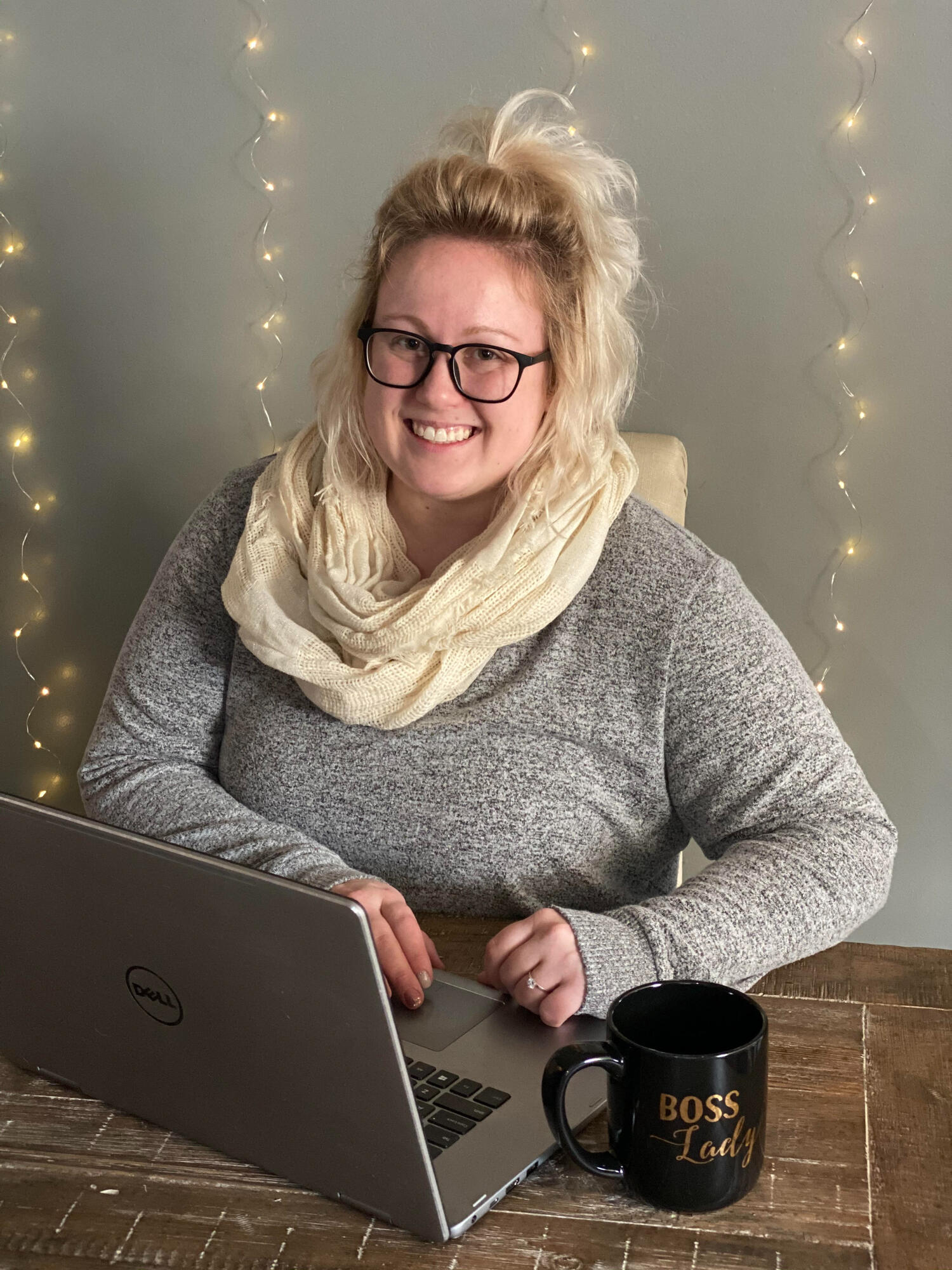 Blonde woman sitting in front of a computer at a table, looking at the camera and smiling.